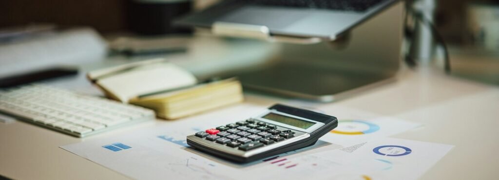 Calculator, notebook and financial charts on a desk in front of a laptop