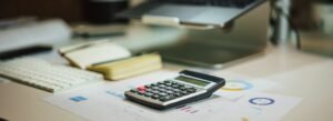 Calculator, notebook and financial charts on a desk in front of a laptop