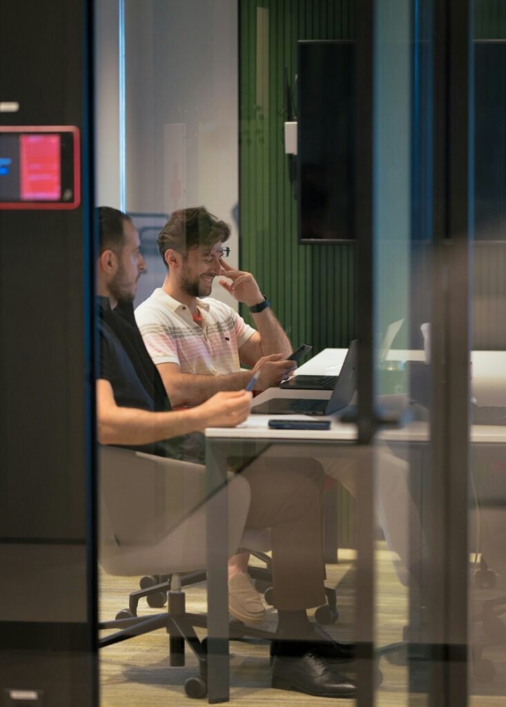 Two men working on laptops at a desk in a glass-walled office
