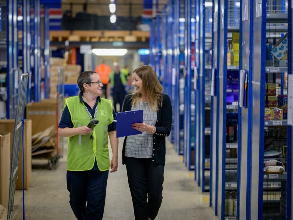 Two women walking through a warehouse aisle with blue shelving units