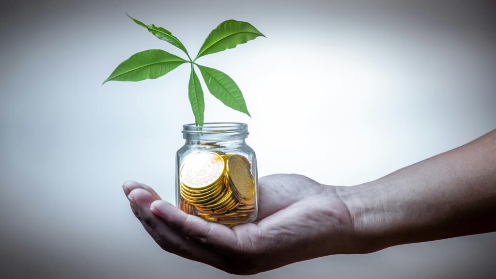 Hand holding a glass jar of gold coins with a young plant growing from it